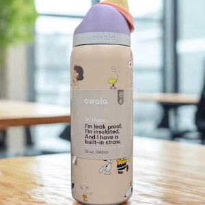 May include: A beige Owala water bottle with a purple lid and a built-in straw. The bottle features cartoon character illustrations and the text "owala". The bottle is 32 oz. / 945ml and has a leak-proof design.