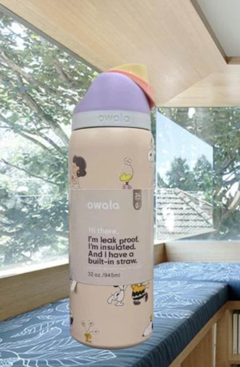 K&ouml;nnte beinhalten: Eine beige Owala-Trinkflasche mit lila Deckel und bunten Akzenten. Die Flasche zeigt Cartoon-Illustrationen und den Text "Hi there. I'm leak proof. I'm insulated. And I have a built-in straw." Fassungsverm&ouml;gen: 945 ml.