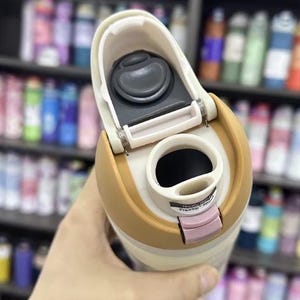 K&ouml;nnte beinhalten: Ein beigefarbener und hellbrauner isolierter Reisebecher mit offenem Deckel. Der Deckel hat einen schwarzen Knopf und einen rosa Verschluss. Der Becher wird in der Hand einer Person gehalten, mit einem verschwommenen Hintergrund von Regalen voller Flaschen.