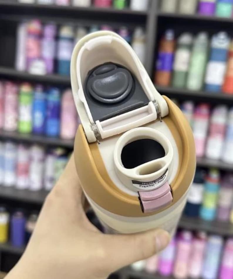 K&ouml;nnte beinhalten: Ein beigefarbener und hellbrauner isolierter Reisebecher mit offenem Deckel. Der Deckel hat einen schwarzen Knopf und einen rosa Verschluss. Der Becher wird in der Hand einer Person gehalten, mit einem verschwommenen Hintergrund von Regalen voller Flaschen.