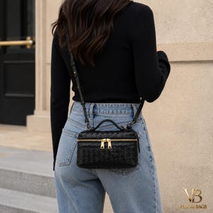 Puede incluir: Bolso bandolera negro de cuero tejido con cremallera y herrajes dorados. El bolso tiene un asa superior y una correa larga ajustable. El texto "VB VELORI BAGS" está en la esquina inferior derecha.