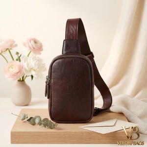 Könnte beinhalten: Eine dunkelbraune Leder-Umhängetasche mit Reißverschluss-Fronttasche und verstellbarem Riemen. Die Tasche wird auf einer Holzoberfläche mit einer Vase mit rosa Blumen und einem Zweig Grünzeug präsentiert. Der Markenname "VELORI BAGS" ist sichtbar.