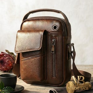 Könnte beinhalten: Braune Leder-Umhängetasche mit oberem Griff und Frontklappentasche. Die Tasche hat ein Reißverschluss-Hauptfach und einen verstellbaren Schultergurt. Metallringdetail und Metallreißverschluss. Die Tasche ist im Satchel-Stil.
