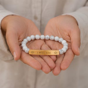 Puede incluir: Una pulsera elástica con cuentas blancas de patrón marmolado y una barra rectangular de metal dorado. La barra tiene grabado el texto I WILL RISE y un pequeño icono de flor de loto a cada lado de la frase, centrado entre las cuentas.