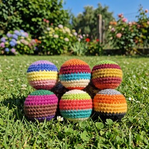 May include: Six colorful, handmade crochet balls arranged on green grass. Each ball features a unique combination of vibrant colors, including blue, yellow, orange, red, green, and purple. The balls are round and appear to be filled with a soft material.