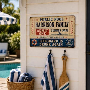 Op de afbeelding: Een rechthoekig metalen bord met een crèmekleurige achtergrond, blauwe en rode tekst en afbeeldingen. Er staat PUBLIC POOL, HARRISON FAMILY, ADMIT ONE, SUMMER PASS, SWIM AT YOUR OWN RISK, LIFEGUARD IS DRUNK AGAIN en VALID ALL SUMMER op, met 000174.