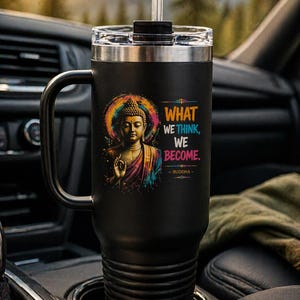 May include: Black travel mug with a handle, featuring a colorful illustration of Buddha with the quote "WHAT WE THINK, WE BECOME." The mug has a clear lid and a straw. The mug is in a car cup holder.
