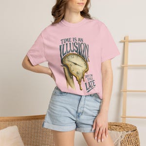 May include: Pink t-shirt with the text "Time is an Illusion" and a melting clock graphic. The clock has the numbers 12, 3, 6, and 9 visible. The text "But I'm Still Late" is below the clock.
