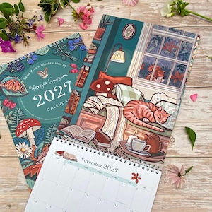 May include: A 2027 wall calendar with spiral binding featuring hand-drawn illustrations by Angie Spurgeon. The cover displays a butterfly, mushrooms, and berries. An open page shows a cozy room with a sleeping orange cat, books, and a tea set.