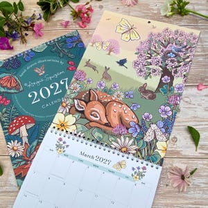 May include: A 2027 wall calendar featuring hand-drawn illustrations by Angie Spurgeon. The cover displays a dark teal background with mushrooms and butterflies. The open page shows a fawn resting in a meadow with rabbits, birds, and flowers.
