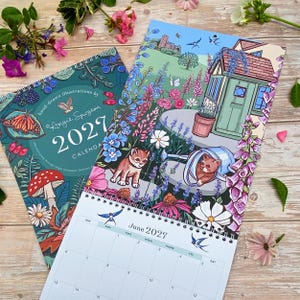 May include: A 2027 wall calendar featuring hand-drawn illustrations by Angie Spurgeon. The cover displays a dark teal background with butterflies, mushrooms, and berries. The open page shows a garden scene with a shed, flowers, and kittens.