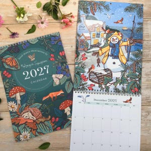 May include: A 2027 wall calendar featuring a dark teal cover with illustrations of a hedgehog, fox, mushrooms, and butterflies, alongside an open page showing a winter scene with a snowman, birds, and a December 2027 grid with numbered dates.