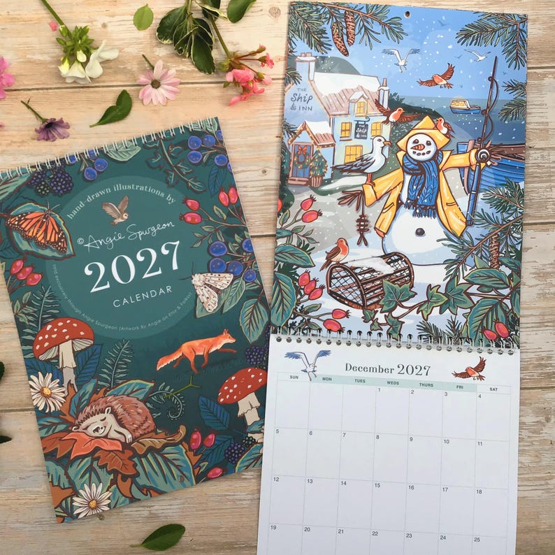 May include: A 2027 wall calendar featuring a dark teal cover with illustrations of a hedgehog, fox, mushrooms, and butterflies, alongside an open page showing a winter scene with a snowman, birds, and a December 2027 grid with numbered dates.