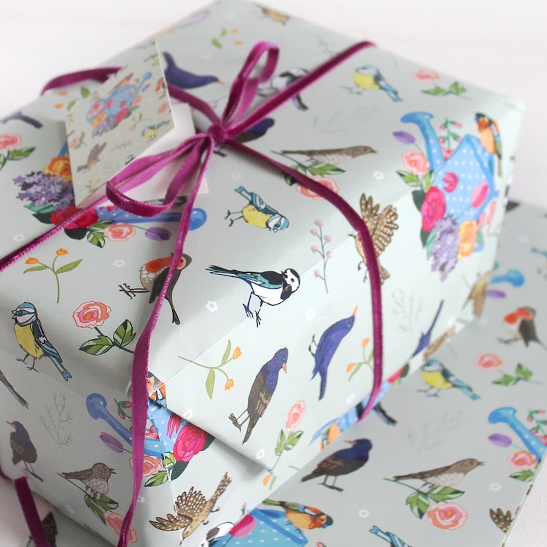 Pu&ograve; includere: Un regalo avvolto in carta da regalo blu e bianca con un motivo di uccelli e fiori. Il regalo &egrave; legato con un nastro rosa.