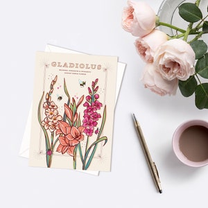 May include: A card with a vintage illustration of gladiolus flowers in shades of pink, peach, and white. The card has the text "Gladiolus" in a retro font and the words "Meaning: Strength & Integrity, August Birth Flower".