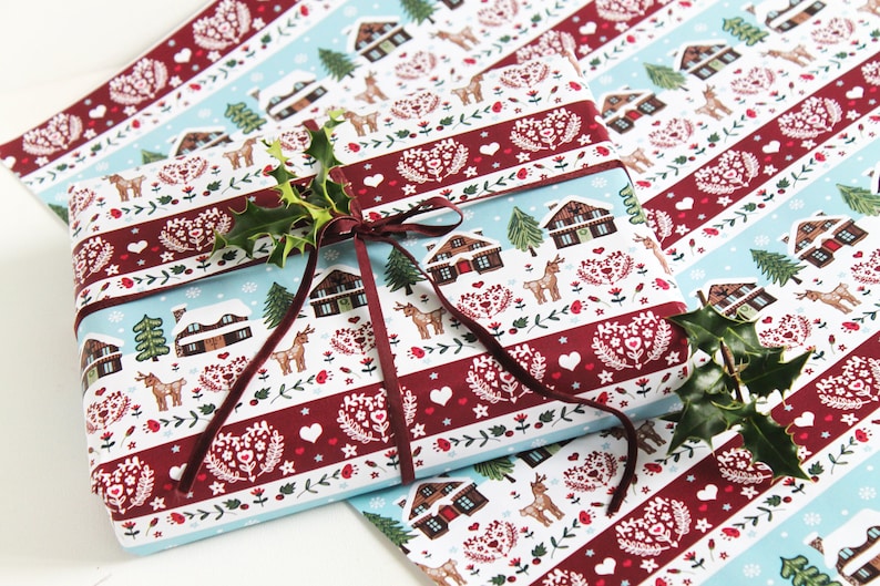 May include: A gift wrapped in blue and red patterned wrapping paper with a brown ribbon. The paper features a winter scene with houses, trees, and deer. The gift is topped with a sprig of holly.