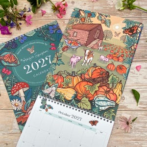 May include: A 2027 spiral-bound wall calendar featuring hand-drawn illustrations by Angie Spurgeon. The cover displays a dark teal background with a butterfly, mushroom, and berries. The open page shows a farm scene with pumpkins, goats, and pigs.