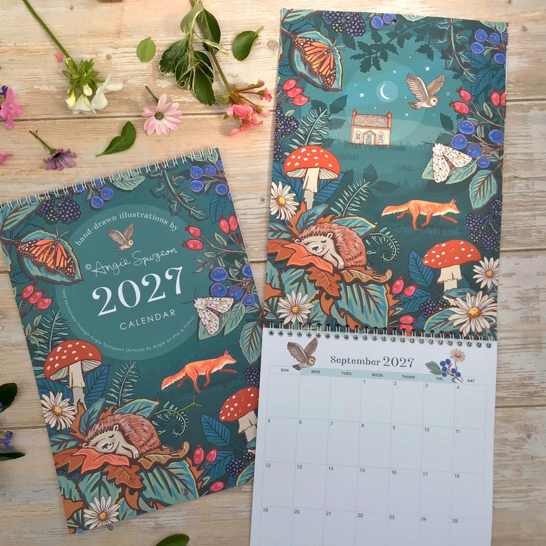 May include: A 2027 wall calendar featuring hand-drawn illustrations by Angie Spurgeon. The cover and pages display a dark teal forest scene with a fox, hedgehog, owl, moth, butterfly, mushrooms, and berries. One page shows the September 2027 grid.