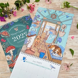 May include: A 2027 wall calendar featuring hand-drawn illustrations by Angie Spurgeon. The cover displays botanical art with a butterfly and mushroom, while the open page shows a beach hut scene with two dogs, a deck chair, and a July 2027 grid.