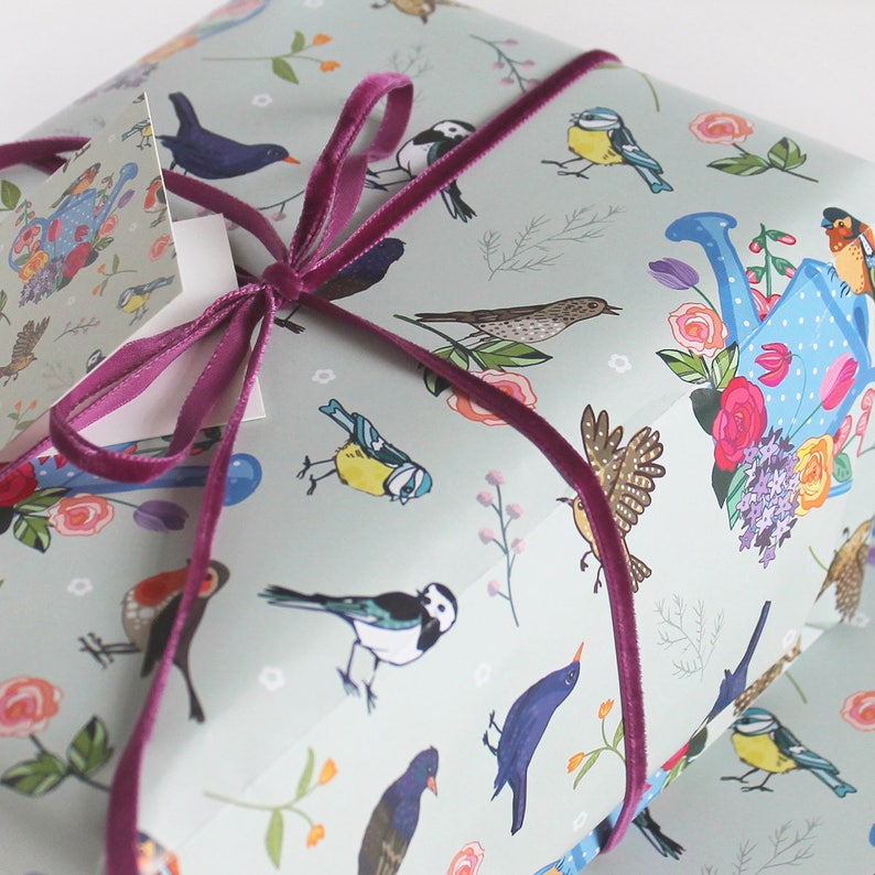 Pu&ograve; includere: Carta da regalo blu chiaro con un motivo ripetitivo di uccelli colorati, fiori e un annaffiatoio. La carta &egrave; legata con un nastro di velluto viola.