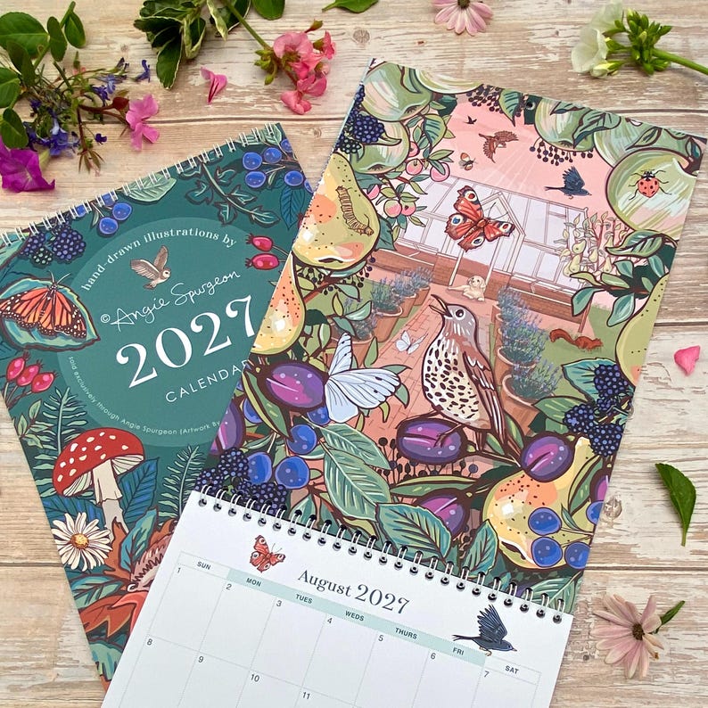 May include: Two 2027 wall calendars with spiral bindings featuring hand-drawn illustrations by Angie Spurgeon. The designs showcase vibrant botanical and wildlife scenes including mushrooms, butterflies, birds, berries, and a greenhouse interior.