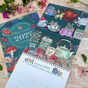 May include: A 2027 wall calendar with a spiral binding featuring hand-drawn illustrations by Angie Spurgeon. The cover displays a butterfly, mushroom, and foliage. The open page shows a February 2027 grid with tea-themed artwork including teapots, cups, and labeled tea tins on shelves.