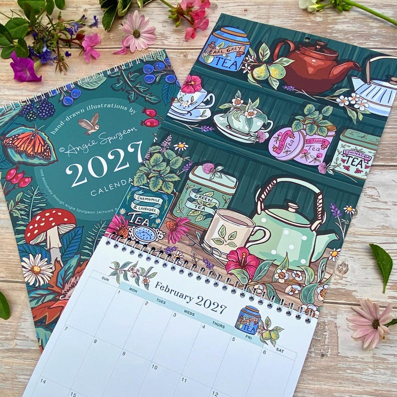 May include: A 2027 wall calendar with a spiral binding featuring hand-drawn illustrations by Angie Spurgeon. The cover displays a butterfly, mushroom, and foliage. The open page shows a February 2027 grid with tea-themed artwork including teapots, cups, and labeled tea tins on shelves.