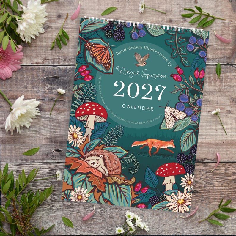 May include: A 2027 wall calendar featuring hand-drawn illustrations by Angie Spurgeon. The cover displays a dark teal background with a monarch butterfly, an owl, a moth, a fox, a hedgehog, mushrooms, berries, and flowers in vibrant colours.