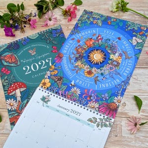 May include: A 2027 wall calendar with hand-drawn illustrations by Angie Spurgeon. The cover features a teal background with a butterfly, mushroom, and berries. An open page shows a blue wheel of the year with seasonal festivals and a January grid.