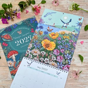 May include: Two 2027 wall calendars with spiral binding. One cover features a dark teal background with a mushroom, butterfly, and owl illustration. The other shows a coastal scene with a puffin, lighthouse, and wildflowers. A May 2027 calendar page is visible.