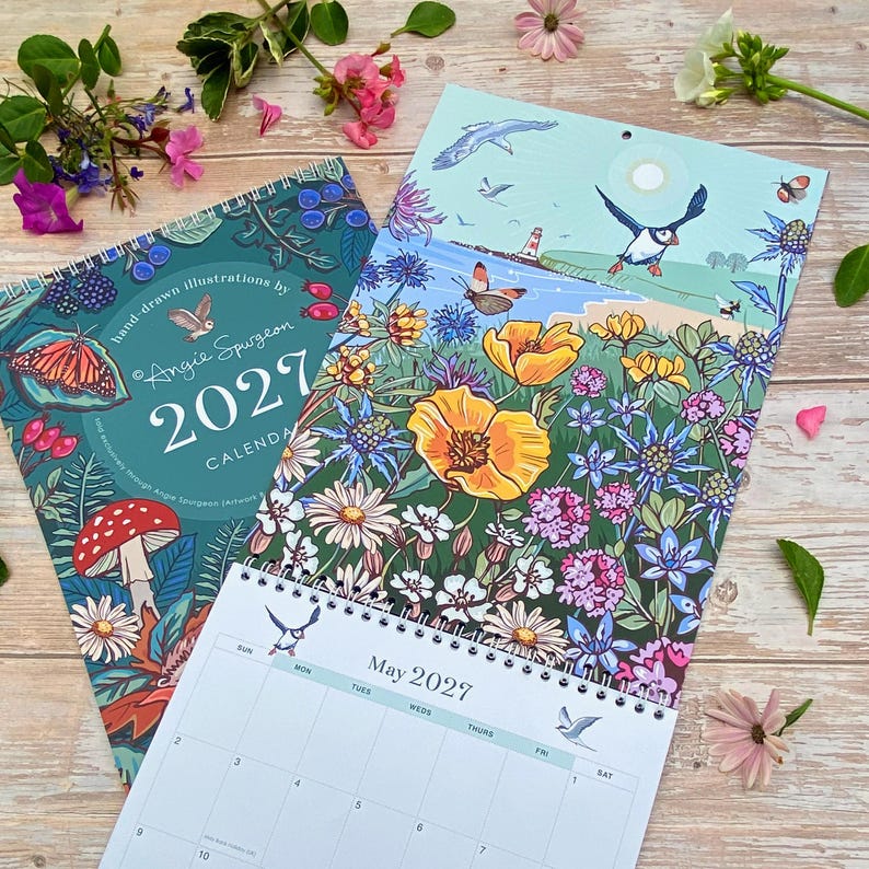 May include: Two 2027 wall calendars with spiral binding. One cover features a dark teal background with a mushroom, butterfly, and owl illustration. The other shows a coastal scene with a puffin, lighthouse, and wildflowers. A May 2027 calendar page is visible.