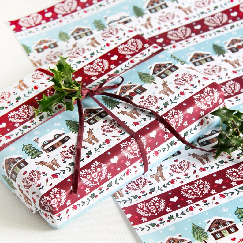 May include: A white and red patterned wrapping paper with a winter village design. The paper features small houses, trees, deer, and hearts. The paper is tied with a burgundy ribbon and decorated with holly.