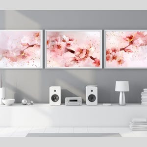 以下が含まれることがあります： 淡いピンクの背景にピンクの桜の枝を描いた水彩画風の長方形のウォールアートプリント3枚組。各プリントは細い白いフレームに収められており、花のディテールが描かれています。