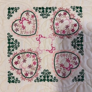 May include: A square off-white fabric panel featuring four embroidered heart motifs in shades of pink and green. Each heart is surrounded by intricate floral and lace-like stitching, with small green corner accents and pink bows in the centre.