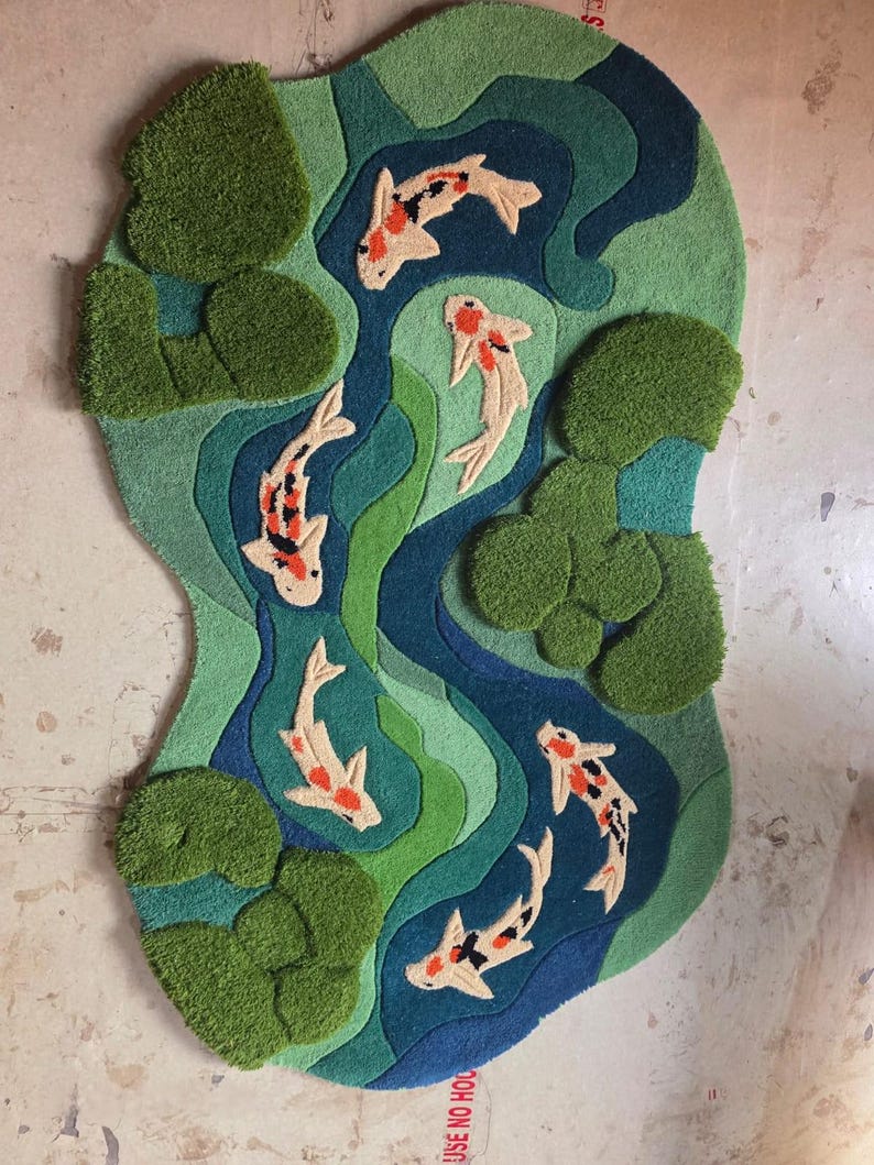 May include: A tufted rug featuring an irregular, organic shape with a pond scene. The design includes shades of green and blue water with several white, orange, and black koi fish swimming among clusters of textured green lily pads.