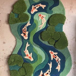 May include: A tufted rug with an irregular shape featuring a pond scene. The design includes five koi fish in white, orange, and black, swimming through layered shades of blue and green water, accented by textured, raised dark green lily pads.