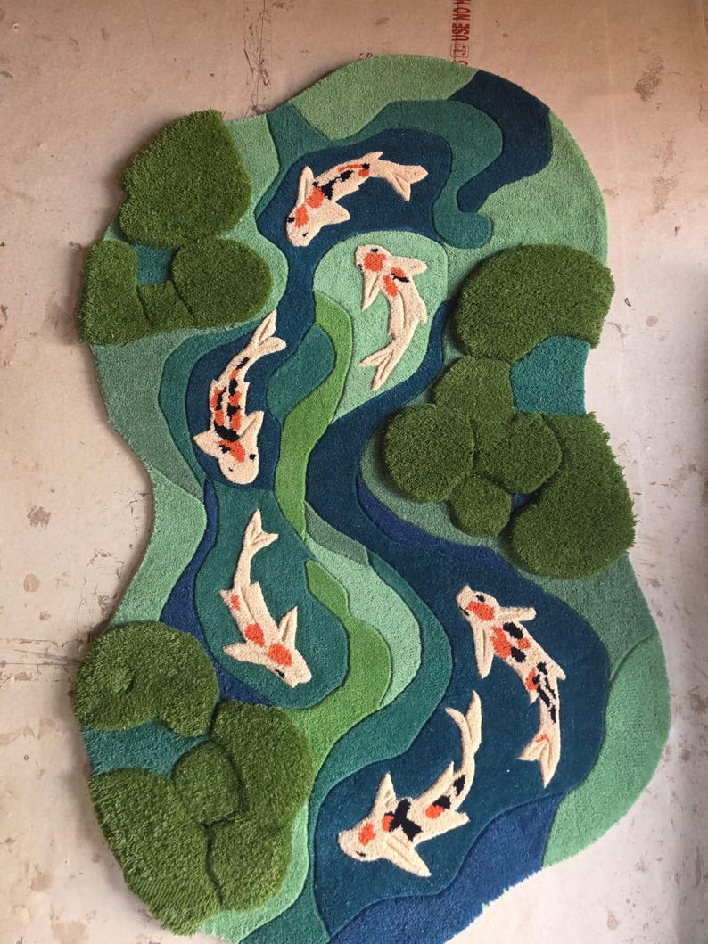 May include: A tufted rug with an irregular shape featuring a pond scene. The design includes five koi fish in white, orange, and black, swimming through layered shades of blue and green water, accented by textured, raised dark green lily pads.