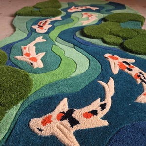 May include: A tufted rug featuring a winding river design in shades of blue and green with raised, textured lily pads and several koi fish in white, orange, and black. The rug has an irregular, organic shape with a high-pile, soft surface.
