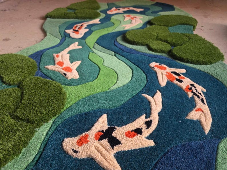 May include: A tufted rug featuring a winding river design in shades of blue and green with raised, textured lily pads and several koi fish in white, orange, and black. The rug has an irregular, organic shape with a high-pile, soft surface.