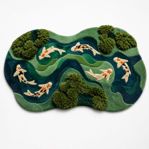 May include: An irregularly shaped tufted rug featuring a pond scene with five koi fish in white, orange, and black. The rug displays layered shades of green and blue to represent water, accented by textured, raised dark green moss-like clusters.
