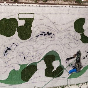 May include: A rectangular tufting frame holding a white fabric backing with a partially completed rug design. The design features outlines of koi fish and abstract green shapes. A blue and silver tufting gun is attached to the fabric.
