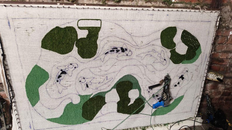 May include: A rectangular tufting frame holding a white fabric backing with a partially completed rug design. The design features outlines of koi fish and abstract green shapes. A blue and silver tufting gun is attached to the fabric.