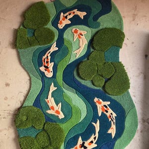 May include: A tufted rug featuring an irregular, organic shape with a pond scene. The design includes six koi fish in white, orange, and black, swimming through layered shades of blue and green water, accented by textured green lily pads.
