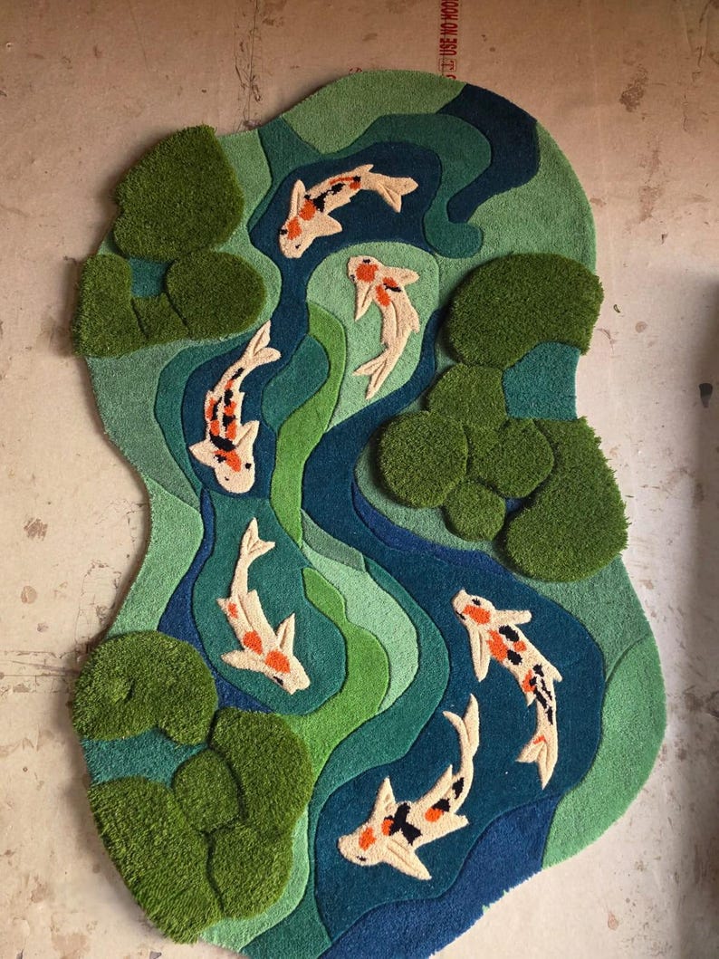 May include: A tufted rug featuring an irregular, organic shape with a pond scene. The design includes six koi fish in white, orange, and black, swimming through layered shades of blue and green water, accented by textured green lily pads.