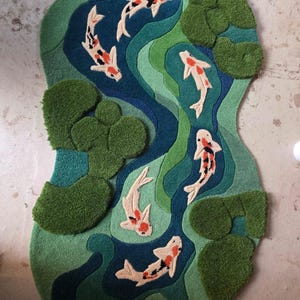 May include: A tufted rug featuring a pond scene with five koi fish in white, orange, and black. The rug has a wavy, organic shape with layered shades of blue and green representing water, accented by textured, raised green lily pads.