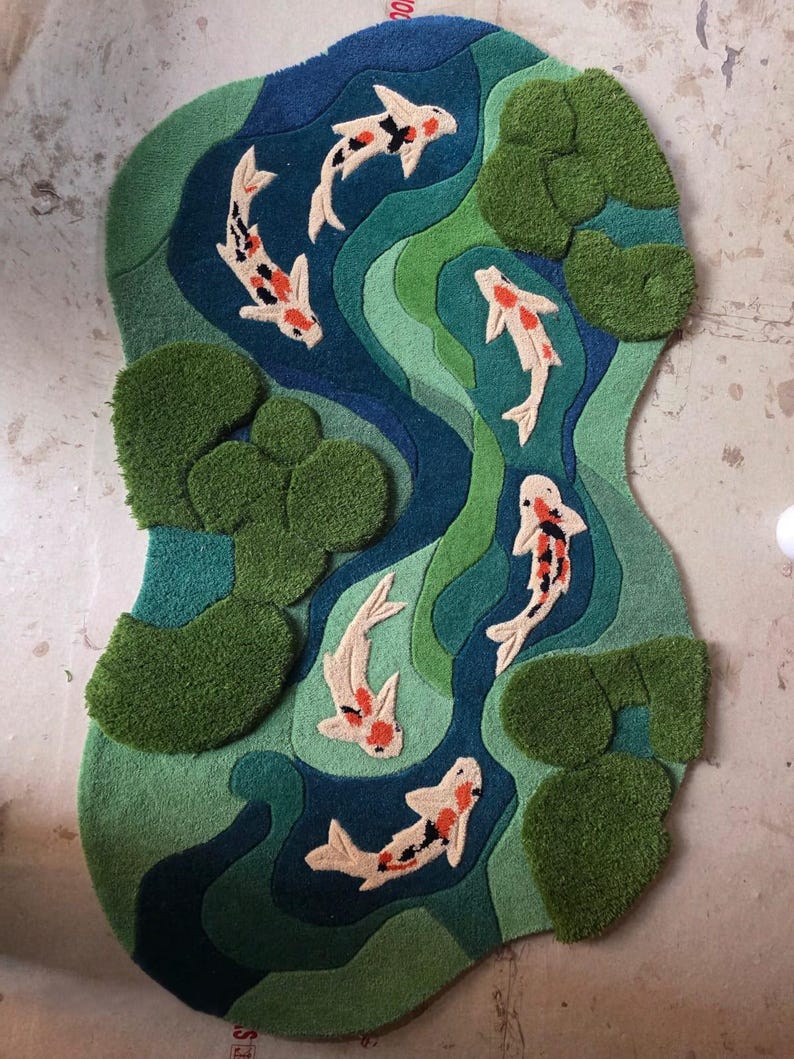 May include: A tufted rug featuring a pond scene with five koi fish in white, orange, and black. The rug has a wavy, organic shape with layered shades of blue and green representing water, accented by textured, raised green lily pads.