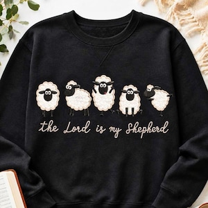 May include: A black crewneck sweatshirt featuring a graphic of five white sheep with black faces and legs. Below the sheep, the text reads "the Lord is my Shepherd" in a cursive, light-coloured font. The garment has long sleeves and a ribbed collar.