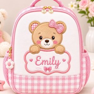 May include: A white and pink gingham backpack featuring a central embroidered brown teddy bear with a pink bow. The bear rests above a white scalloped plaque with the name Emily embroidered in pink, flanked by two small pink hearts and a bow.