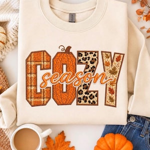 May include: A cream-colored crewneck sweatshirt featuring the word COZY in large block letters with varied patterns including orange plaid, a pumpkin texture, leopard print, and autumn leaves, with the word season in orange cursive script.