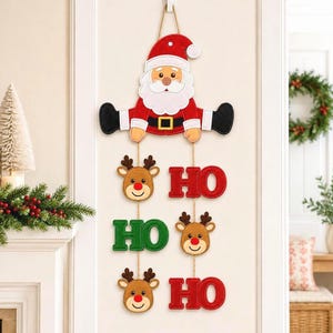 Könnte beinhalten: Eine hängende Weihnachtswanddekoration mit einer Filzfigur des Weihnachtsmanns an der Spitze, gefolgt von drei Rentiergesichtern und drei Schriftzügen HO in Rot und Grün, die mit dünnen Bindfäden verbunden und an einem weißen Haken aufgehängt sind.